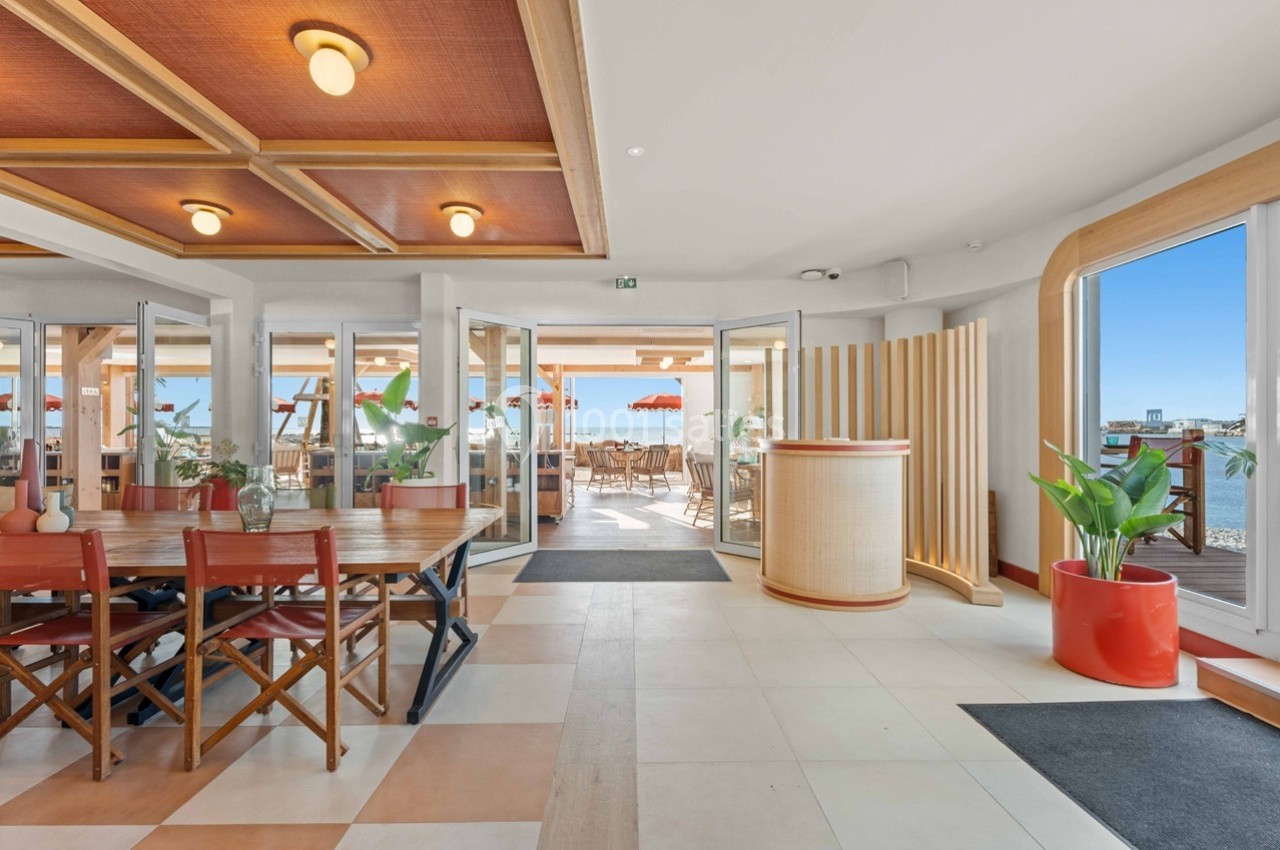 Intérieur lumineux d'un restaurant avec vue sur la mer, mobilier en bois, plantes décoratives et grandes baies vitrées.