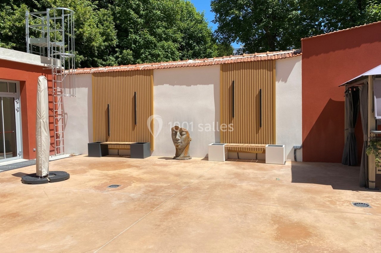 Cour extérieure avec mur blanc et orange, bancs en bois, sculpture de tête et échelle métallique à gauche.