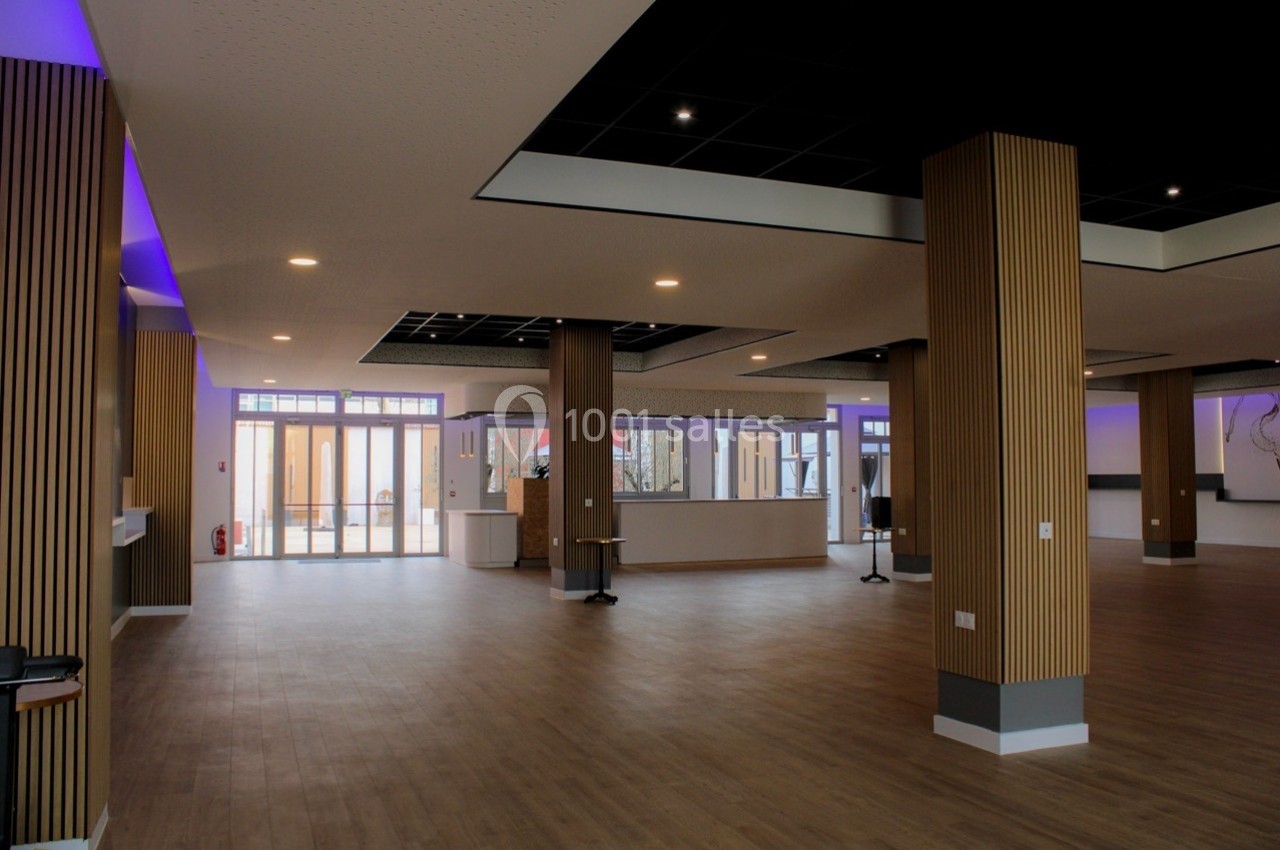 Salle spacieuse avec parquet, colonnes en bois, plafond noir et éclairage violet, donnant sur des baies vitrées.