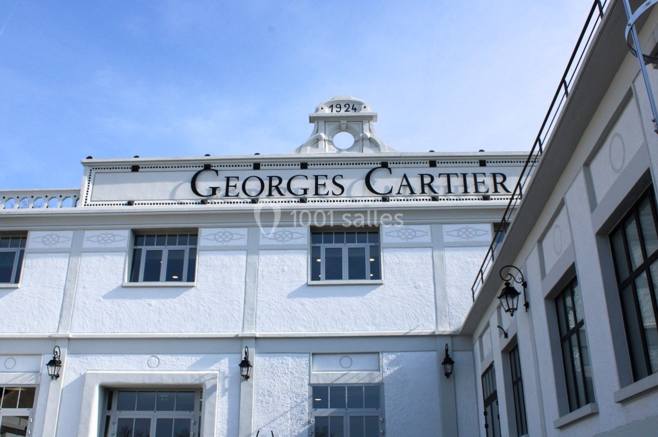 Façade blanche d'un bâtiment avec l'inscription ’Georges Cartier’ et l'année 1924, sous un ciel bleu clair.