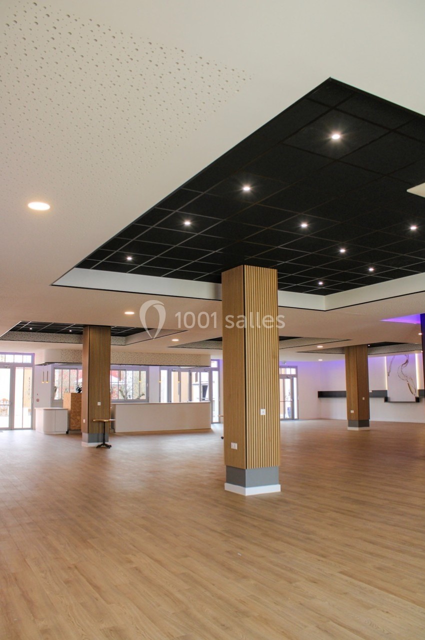 Salle spacieuse avec parquet, colonnes en bois, plafond noir à spots lumineux et grandes baies vitrées.