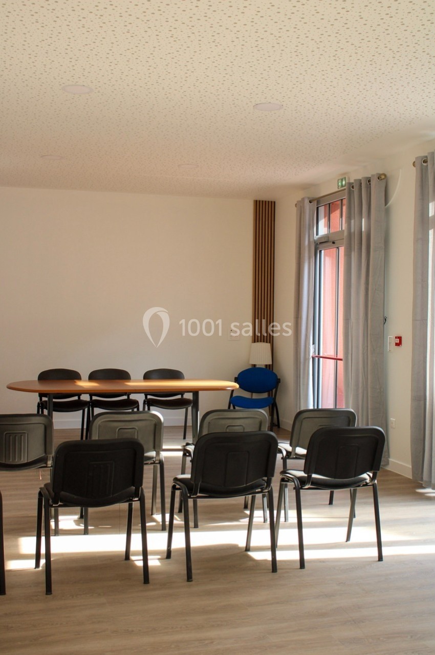 Salle de réunion lumineuse avec des chaises alignées face à une table ovale, près de grandes fenêtres avec rideaux.