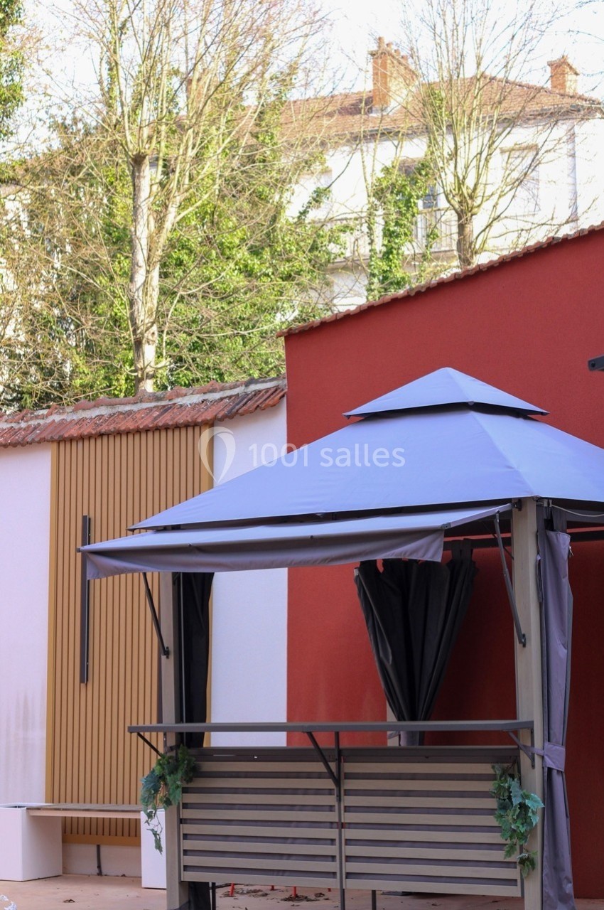 Tonnelle avec rideaux noirs dans une cour, entourée de murs rouges et blancs, avec des arbres en arrière-plan.