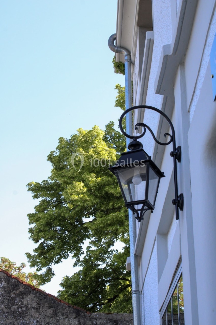 Lampe murale noire fixée sur une façade blanche, avec un arbre et un ciel bleu en arrière-plan.