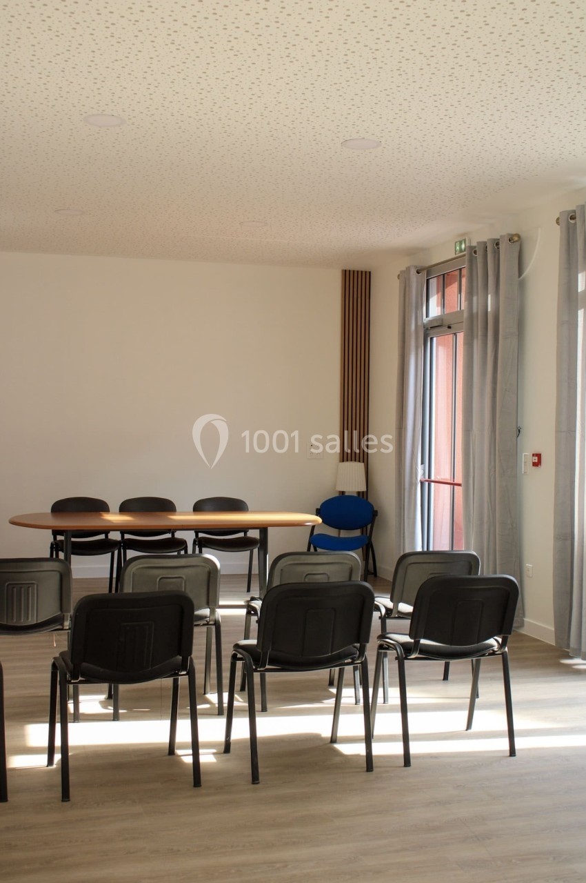 Salle de réunion lumineuse avec des chaises alignées face à une table ovale, près de grandes fenêtres avec rideaux.