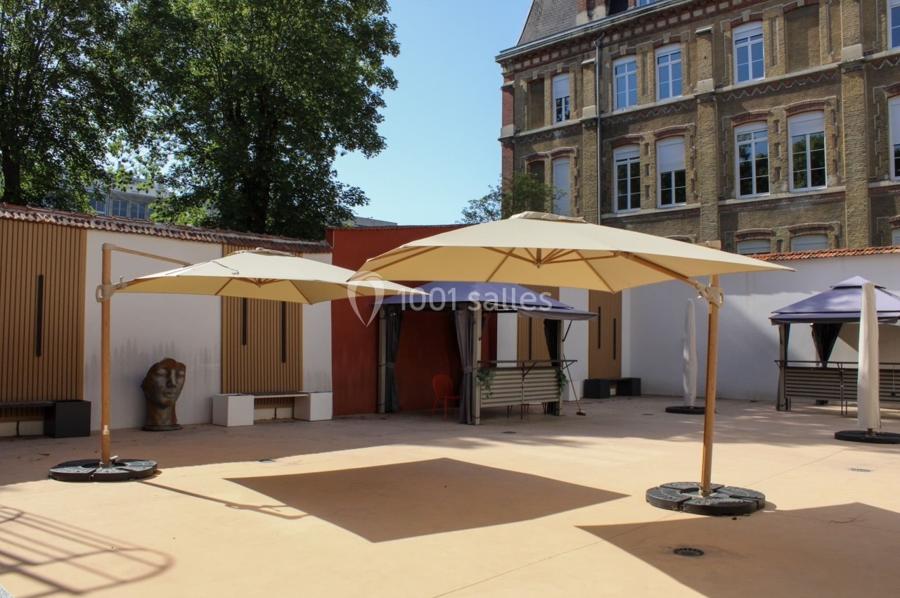 Cour extérieure avec parasols beige, cabines ouvertes, sculpture et bâtiment en briques en arrière-plan.