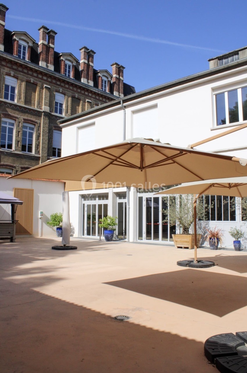 Cour extérieure ensoleillée avec parasols, sol beige, plantes en pots et bâtiments en arrière-plan.
