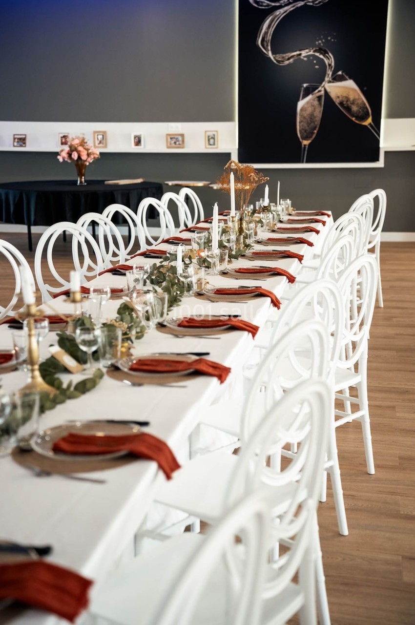 Table dressée avec des chaises blanches, serviettes rouges, bougies et décoration végétale dans une salle élégante.