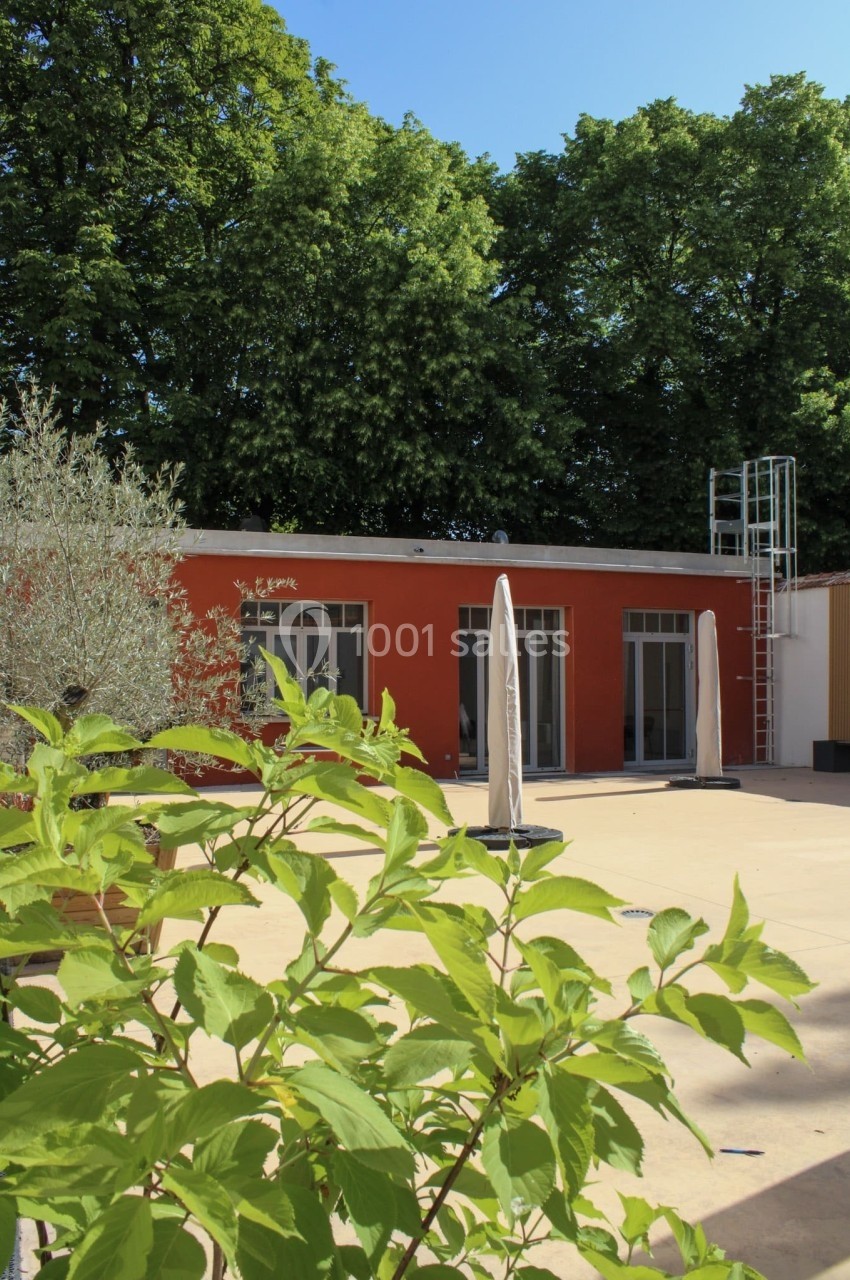 Façade rouge d'un bâtiment avec grandes fenêtres, entouré de végétation et donnant sur une terrasse ensoleillée.