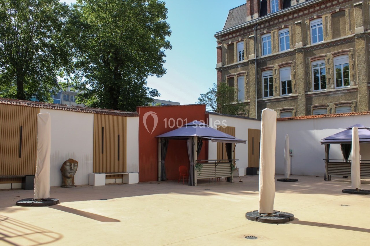 Cour extérieure avec sol en béton, parasols pliés, bancs, et bâtiments en briques avec fenêtres en arrière-plan.
