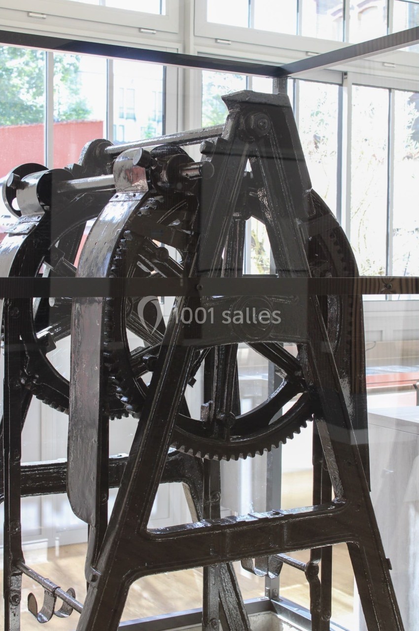 Machine industrielle ancienne en métal noir, exposée dans un espace lumineux avec de grandes fenêtres en arrière-plan.