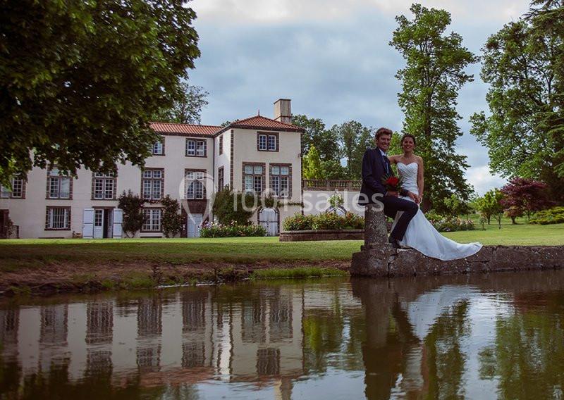 Un couple en tenue de mariage pose près d'un étang devant une grande maison entourée de verdure.
