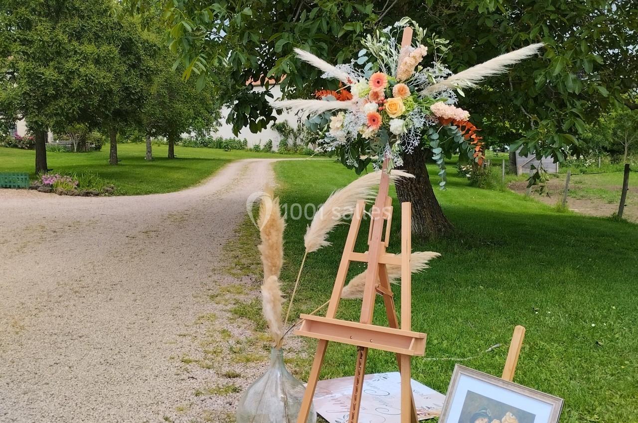 Chevalet en bois avec bouquet floral et tableau, disposé près d'un chemin gravillonné dans un jardin verdoyant.