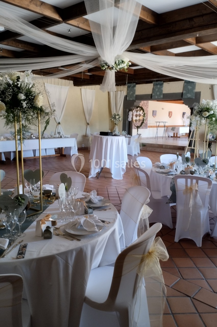 Salle décorée pour un événement avec tables rondes dressées, nappes blanches, chaises ornées et compositions florales.