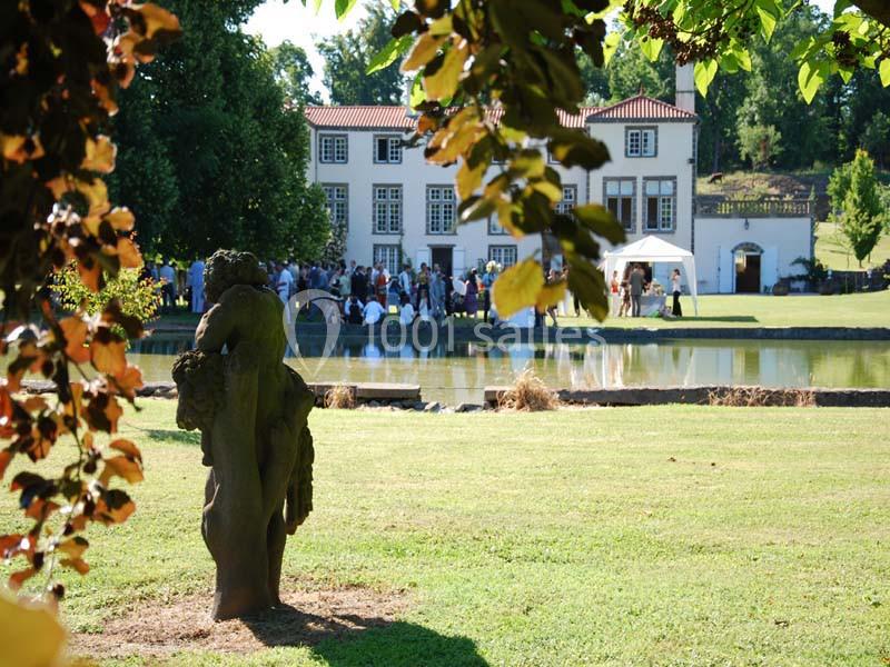 Statue en pierre dans un parc verdoyant, avec un étang et une grande maison en arrière-plan.