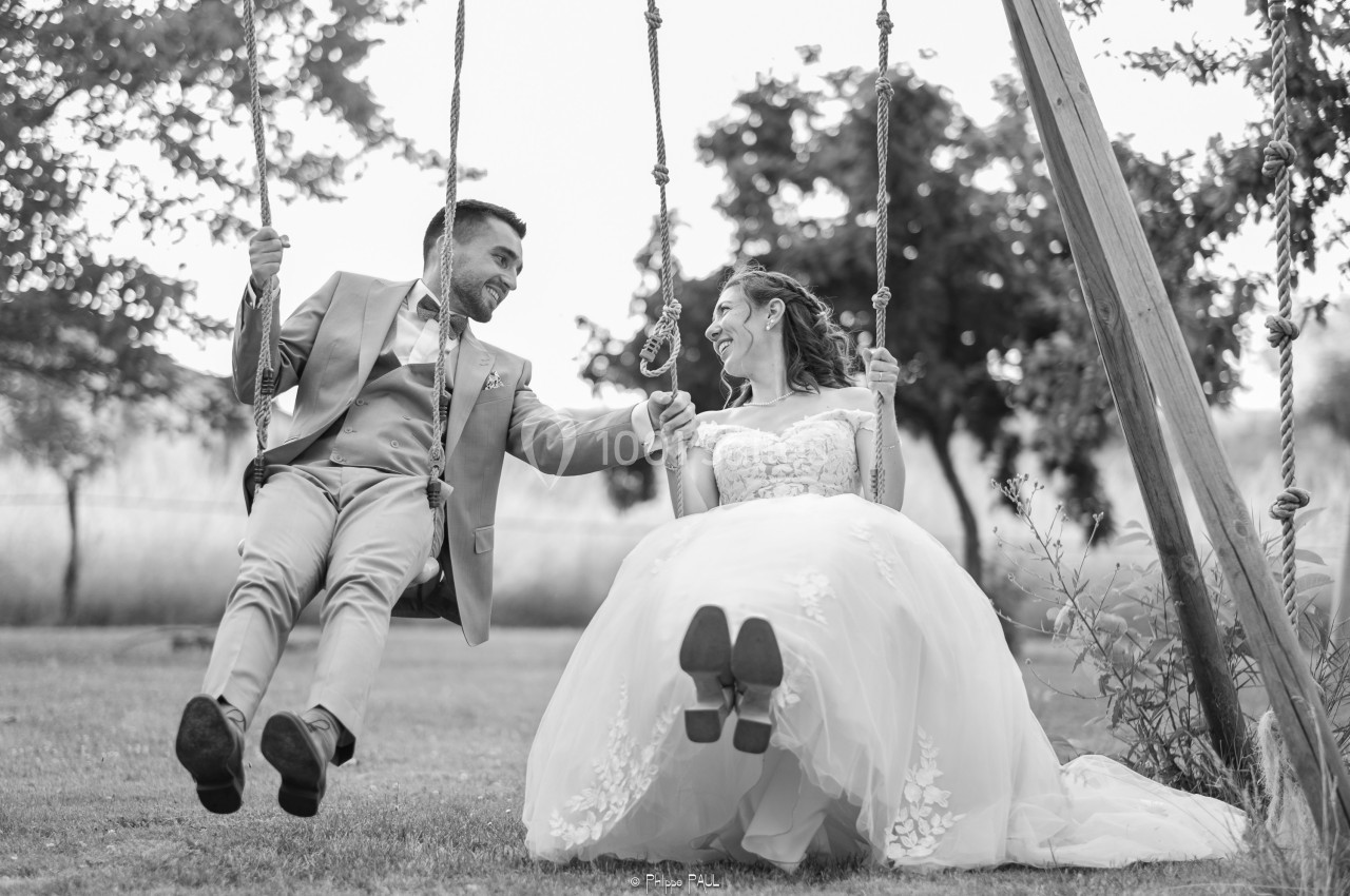 Un couple en tenue de mariage sourit en se balançant côte à côte sur une balançoire dans un jardin.
