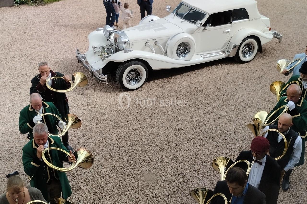 Un groupe de musiciens jouant du cor autour d'une voiture ancienne blanche garée sur une allée gravillonnée.