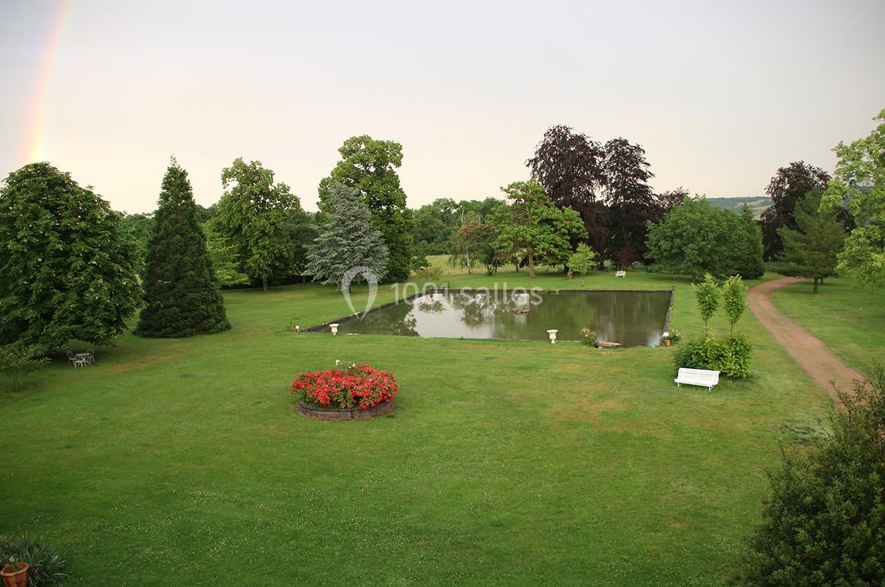 Vue d'un jardin verdoyant avec un étang, des arbres, des fleurs rouges et un chemin en gravier sous un ciel clair.