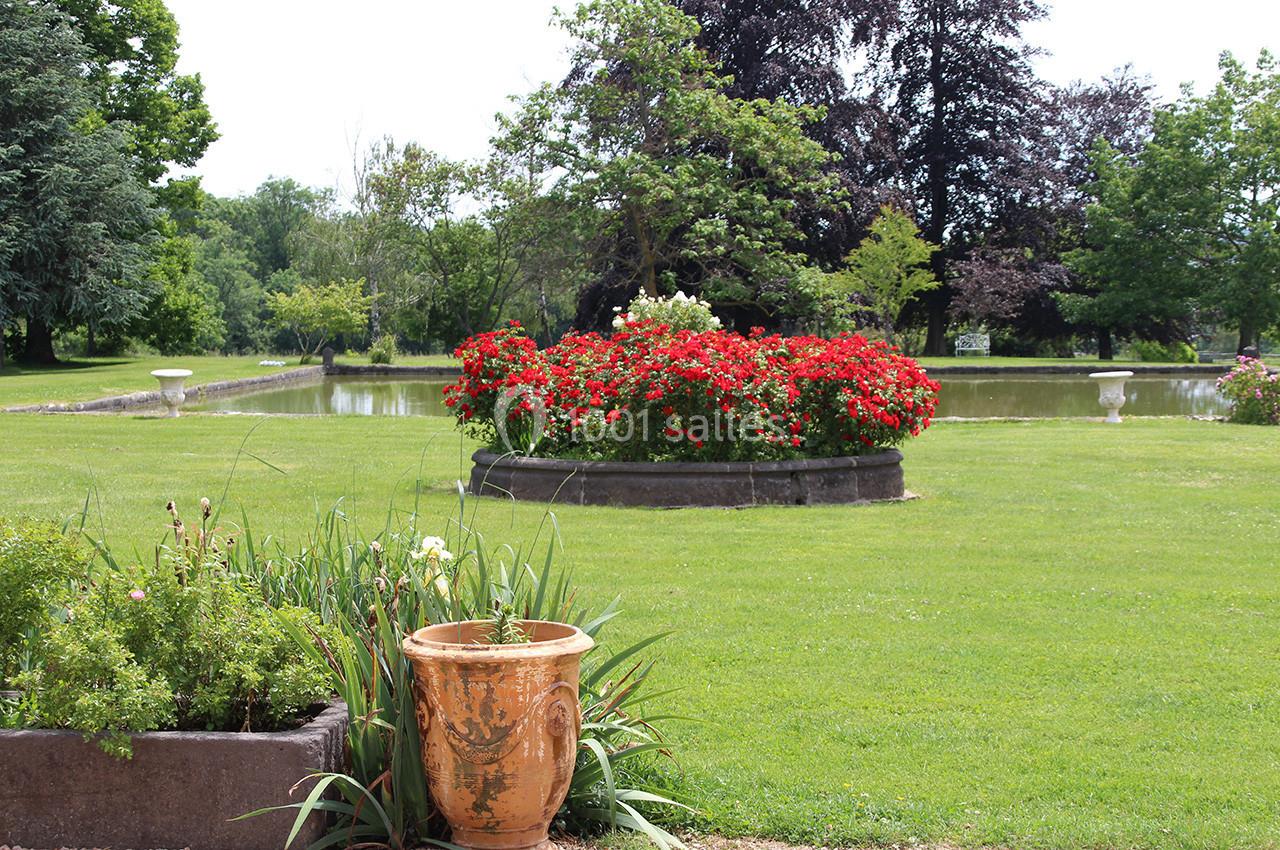 Jardin avec pelouse, massif de fleurs rouges, pot en terre cuite au premier plan et bassin d'eau en arrière-plan.