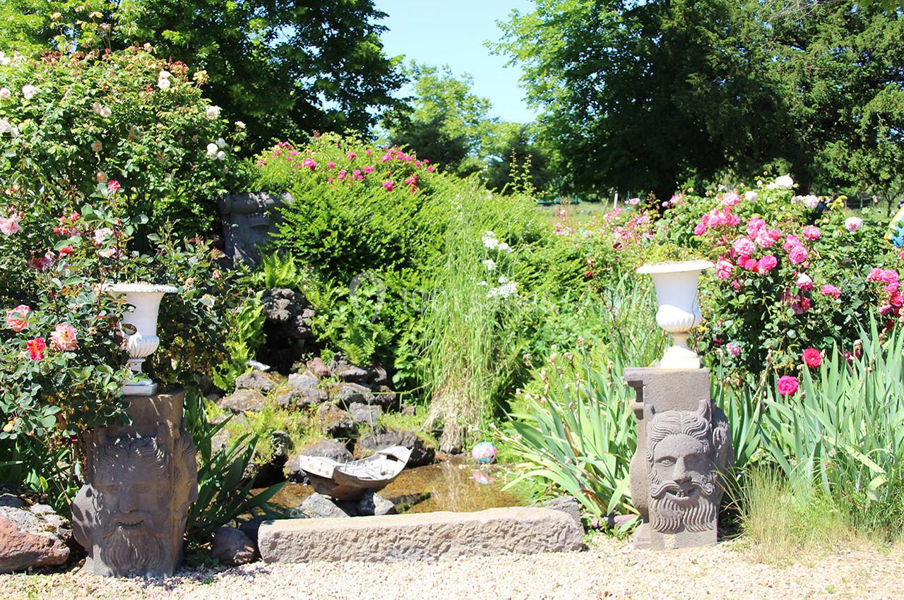 Jardin fleuri avec rosiers, statues en pierre et un petit ruisseau entouré de verdure.
