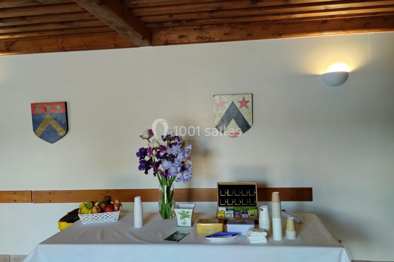 Table dressée avec des fleurs, fruits, boissons et accessoires, dans une salle avec des blasons accrochés au mur.