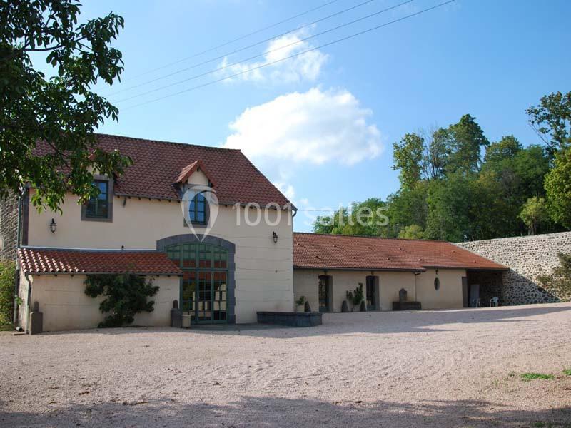 Bâtiment rural rénové avec façade claire, toit en tuiles rouges, entouré d'arbres et d'une cour en gravier.