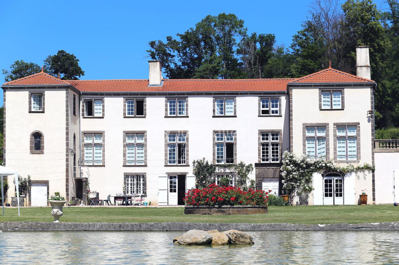 Grande maison blanche à trois étages avec toit en tuiles rouges, entourée de verdure et située près d'un plan d'eau.