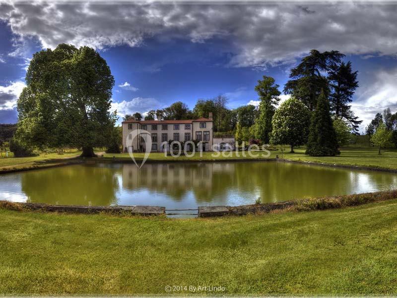 Manoir entouré d'arbres, reflété dans un étang sous un ciel partiellement nuageux.