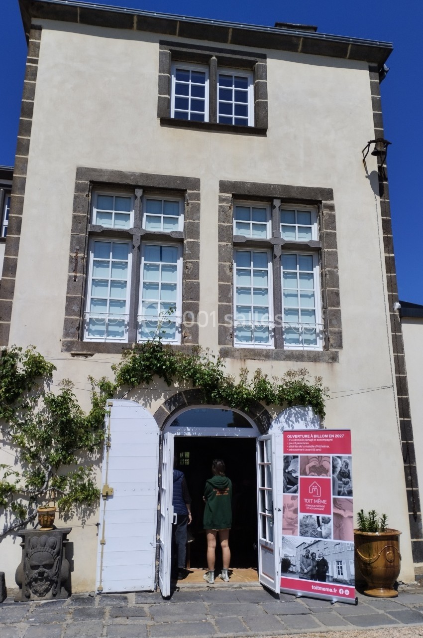 Le gîte Façade d'un bâtiment en pierre avec une porte ouverte, des visiteurs entrant et un panneau d'exposition à droite.
