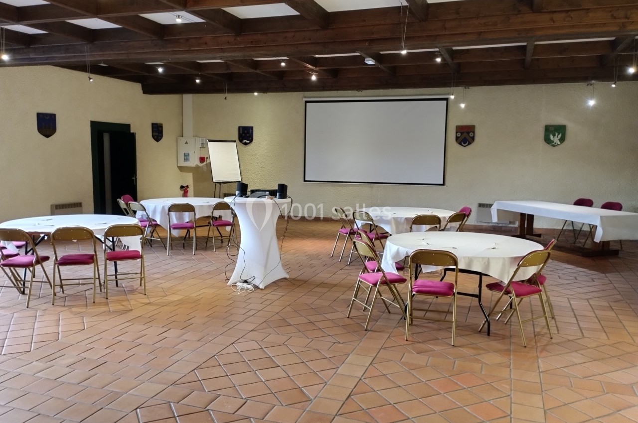 Salle des armoiries Salle de réunion avec tables rondes, chaises pliantes, vidéoprojecteur et écran, dans un espace au plafond en bois.