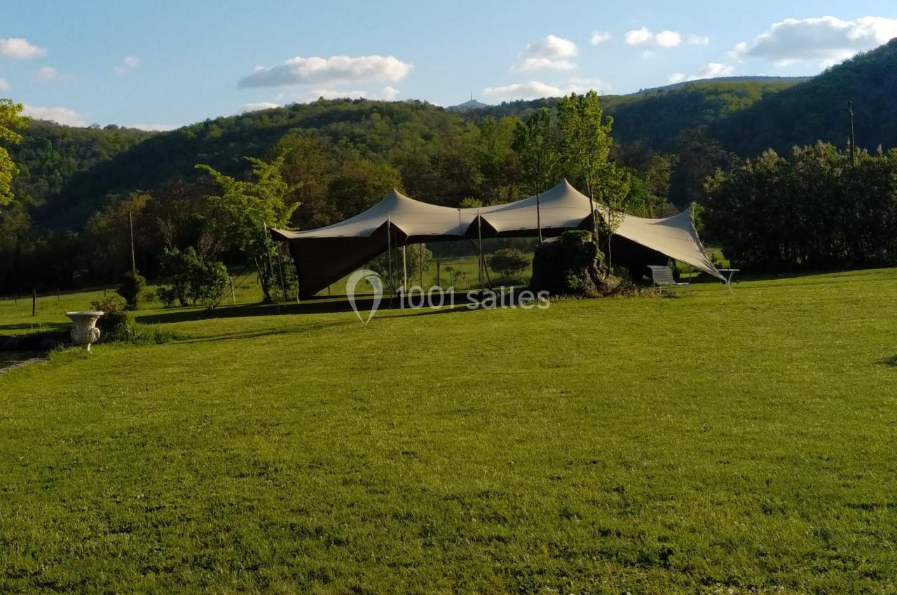 Tente beige installée sur une pelouse verdoyante, entourée de collines et d'arbres sous un ciel partiellement nuageux.