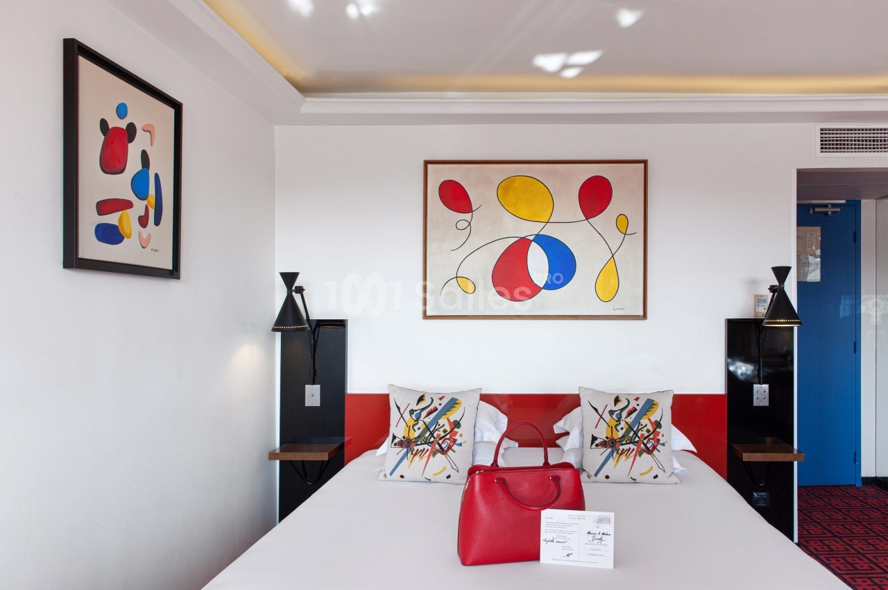 Chambre d'hôtel moderne avec lit double, coussins colorés, tableaux abstraits et sac rouge posé sur le lit.