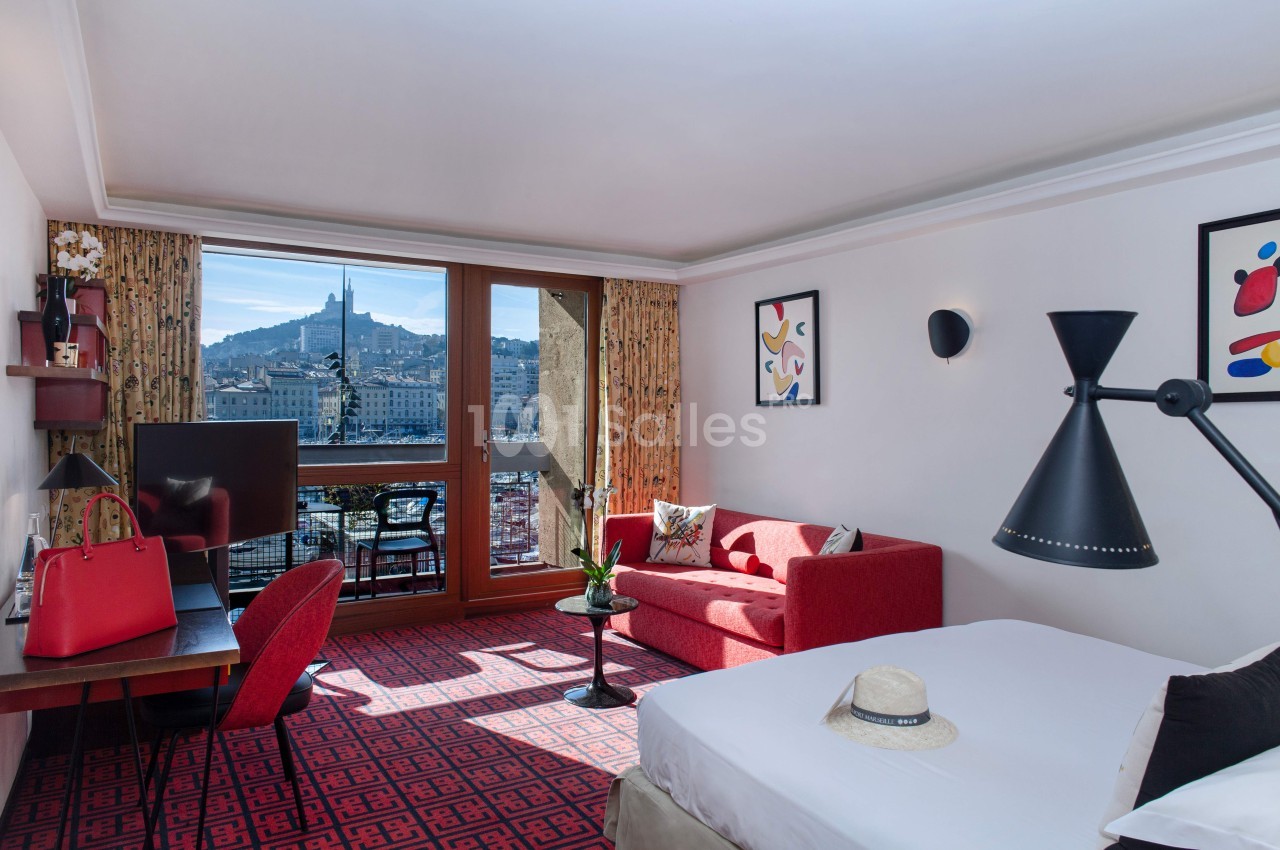 Chambre d'hôtel lumineuse avec vue sur une basilique, canapé rouge, bureau, lit double et balcon aménagé.