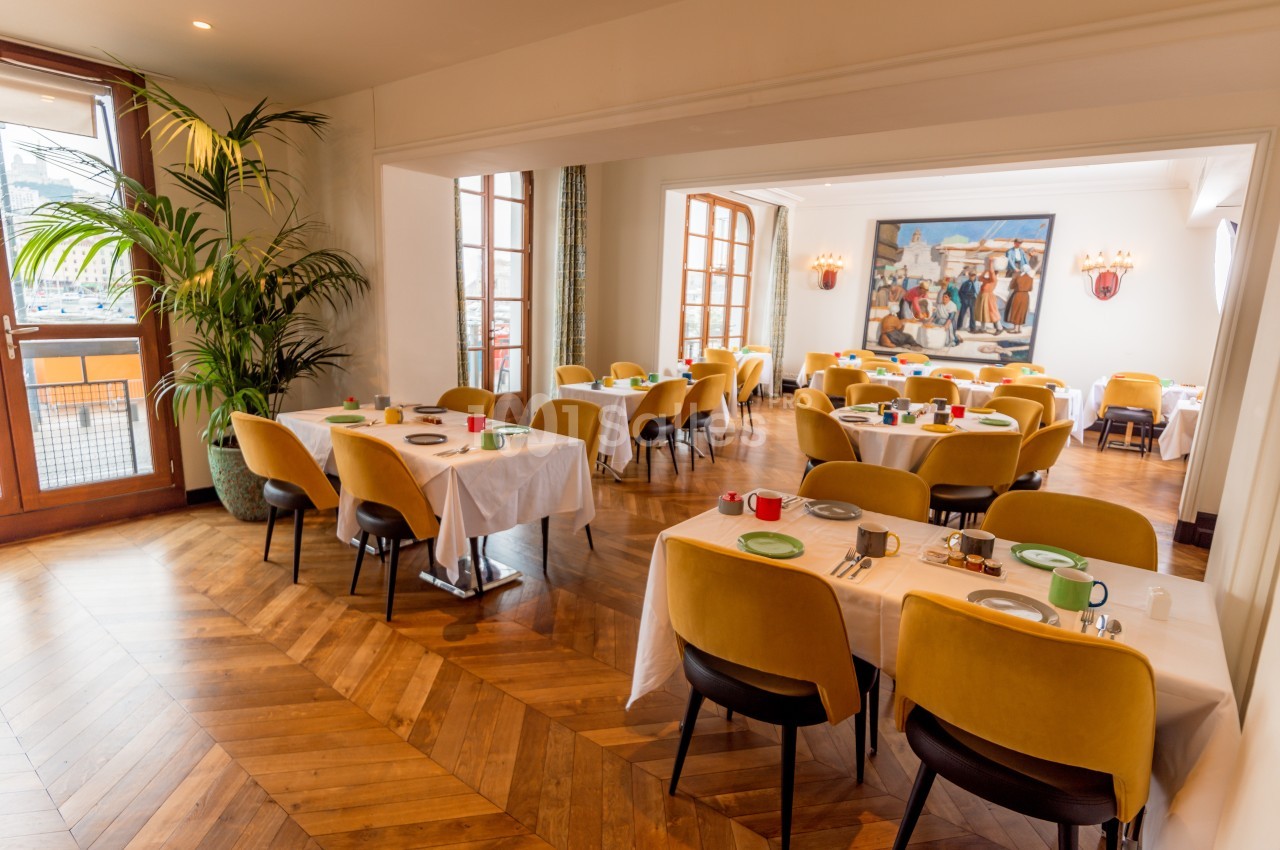 Salle de restaurant lumineuse avec tables dressées, chaises jaunes, parquet en bois et tableau mural coloré.