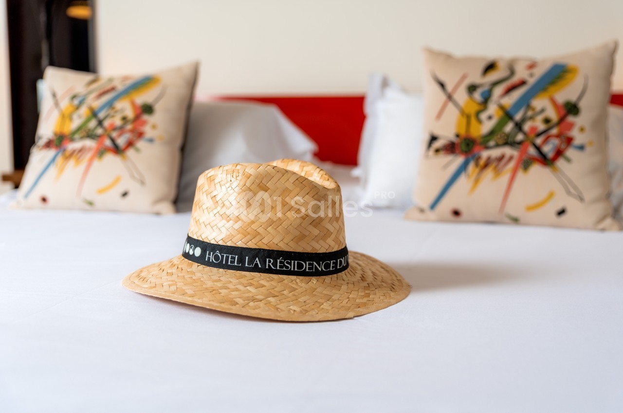 Chapeau de paille avec un ruban marqué ’Hôtel La Résidence’ posé sur un lit blanc, coussins colorés en arrière-plan.