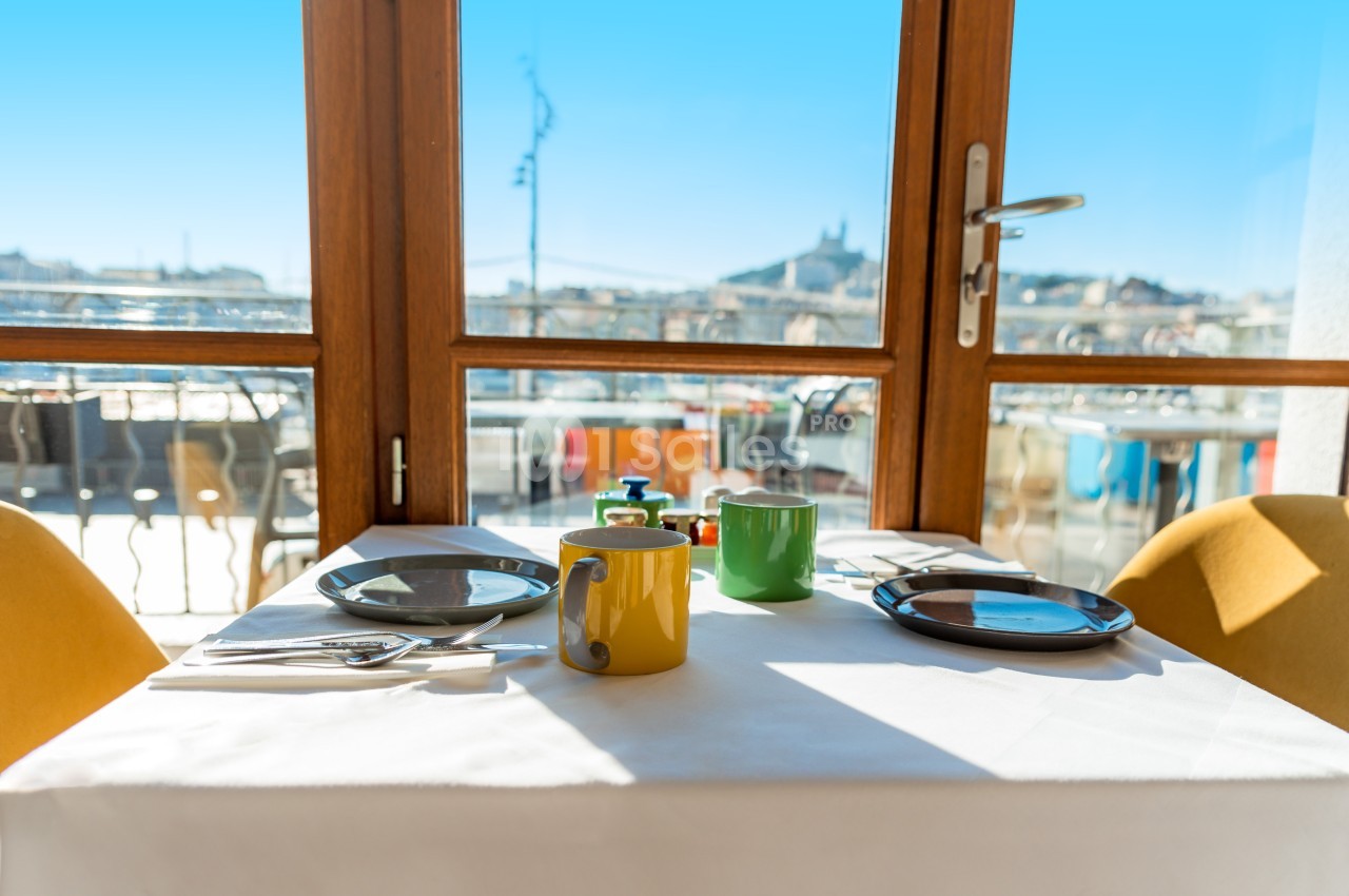 Table dressée avec vaisselle colorée devant une baie vitrée offrant une vue sur une ville ensoleillée.