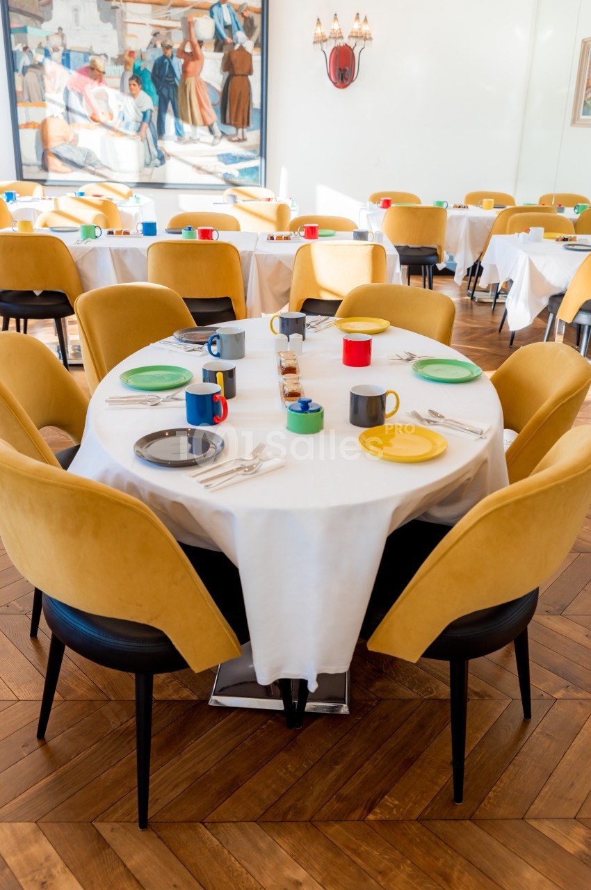 Table dressée avec nappes blanches, assiettes colorées, tasses et couverts, entourée de chaises jaunes dans une salle…