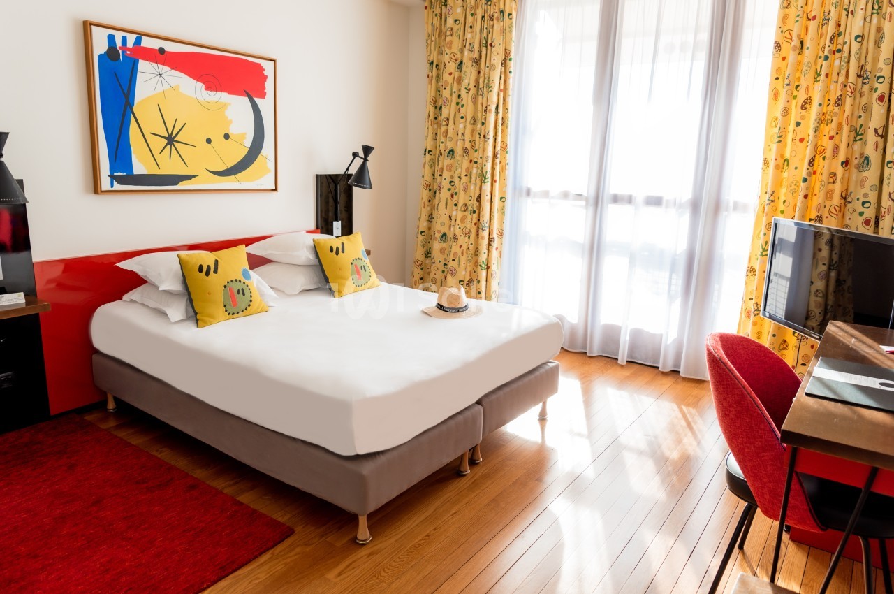 Chambre lumineuse avec lit double, coussins décoratifs, bureau, fauteuil rouge et tableau coloré au mur.
