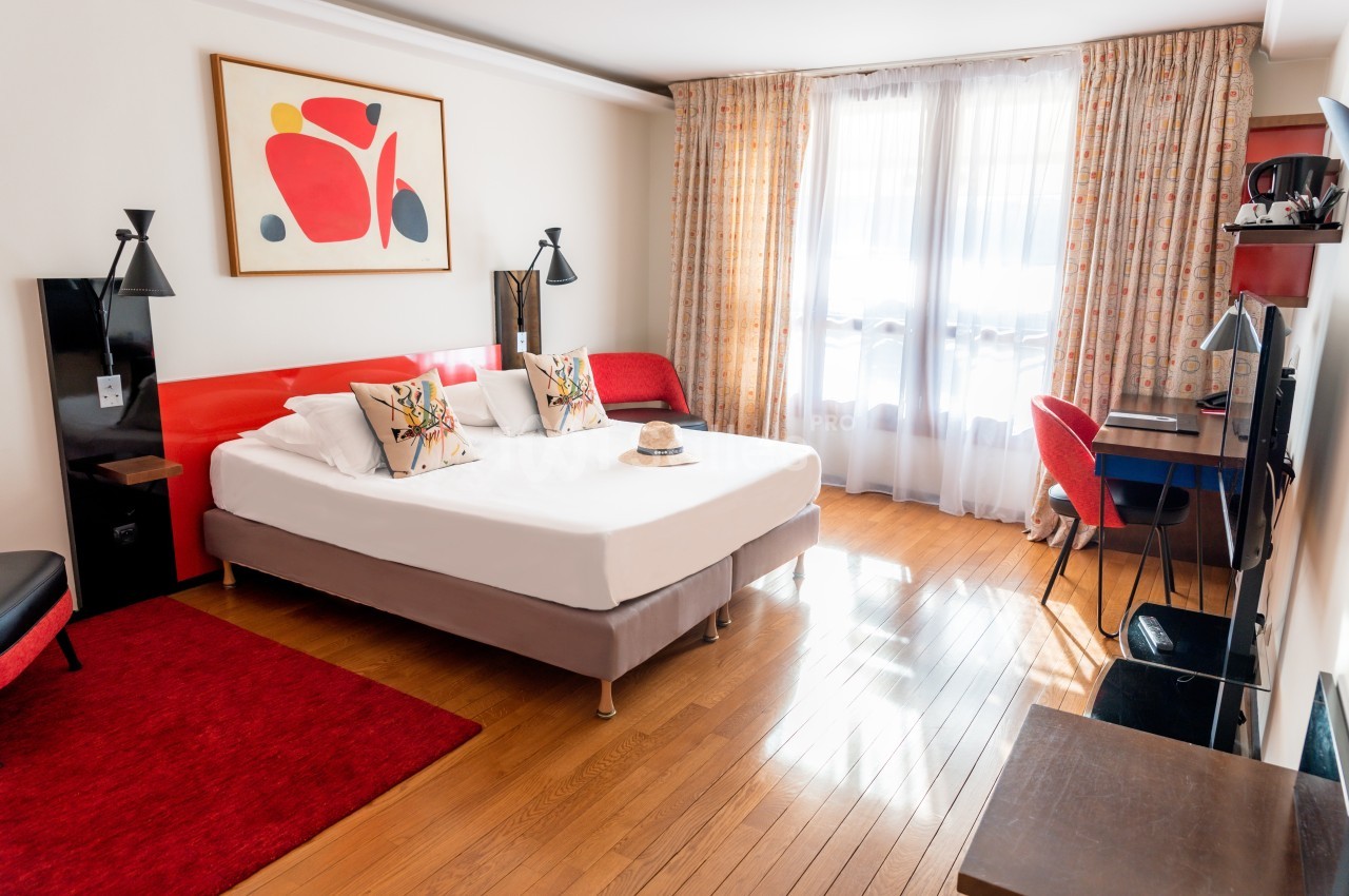 Chambre lumineuse avec lit double, décoration moderne, bureau, fauteuils et grandes fenêtres avec rideaux.