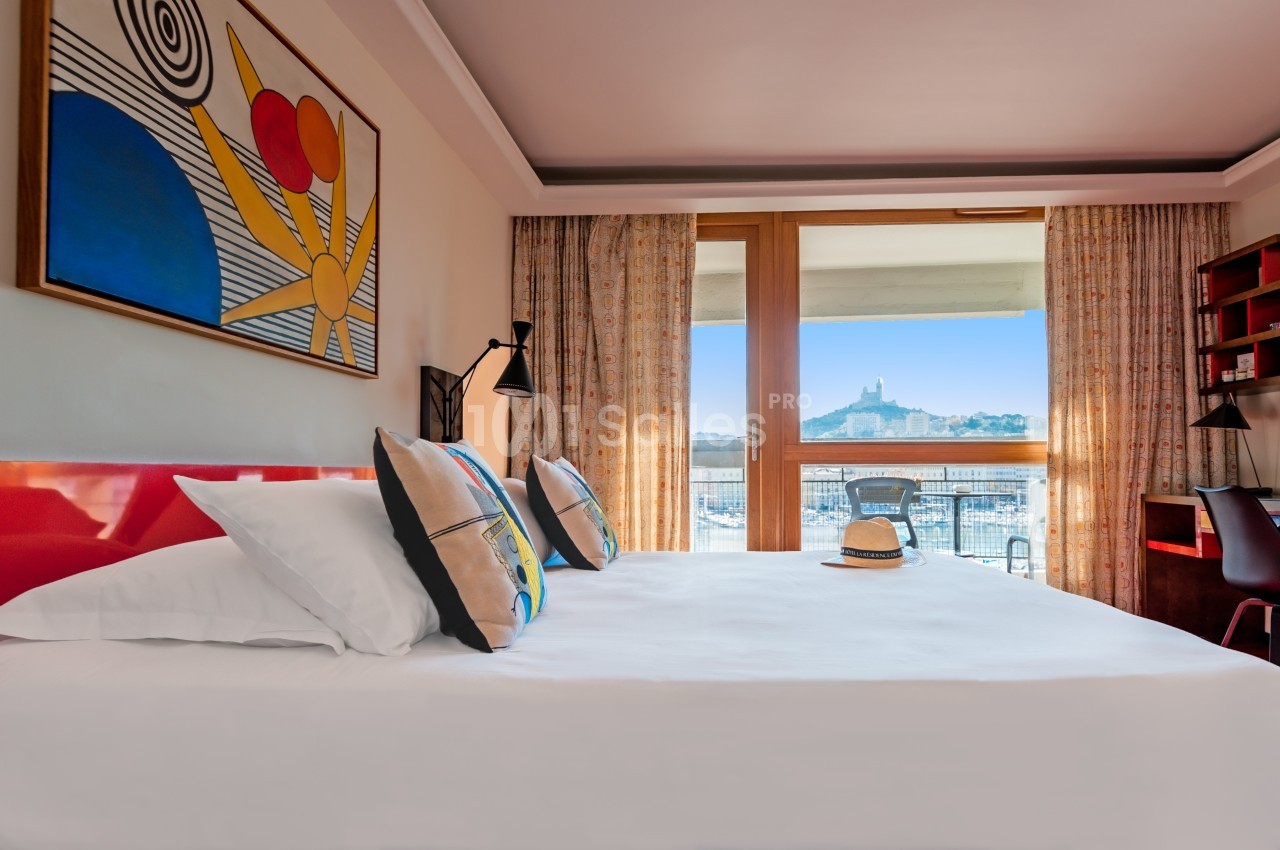 Chambre d'hôtel lumineuse avec lit double, vue sur un port et un monument depuis une grande baie vitrée.