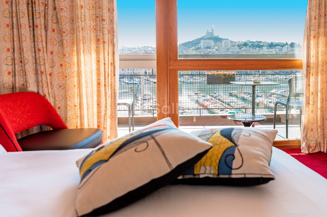 Vue sur le Vieux-Port de Marseille et Notre-Dame de la Garde depuis une chambre d'hôtel avec balcon et fauteuil rouge.