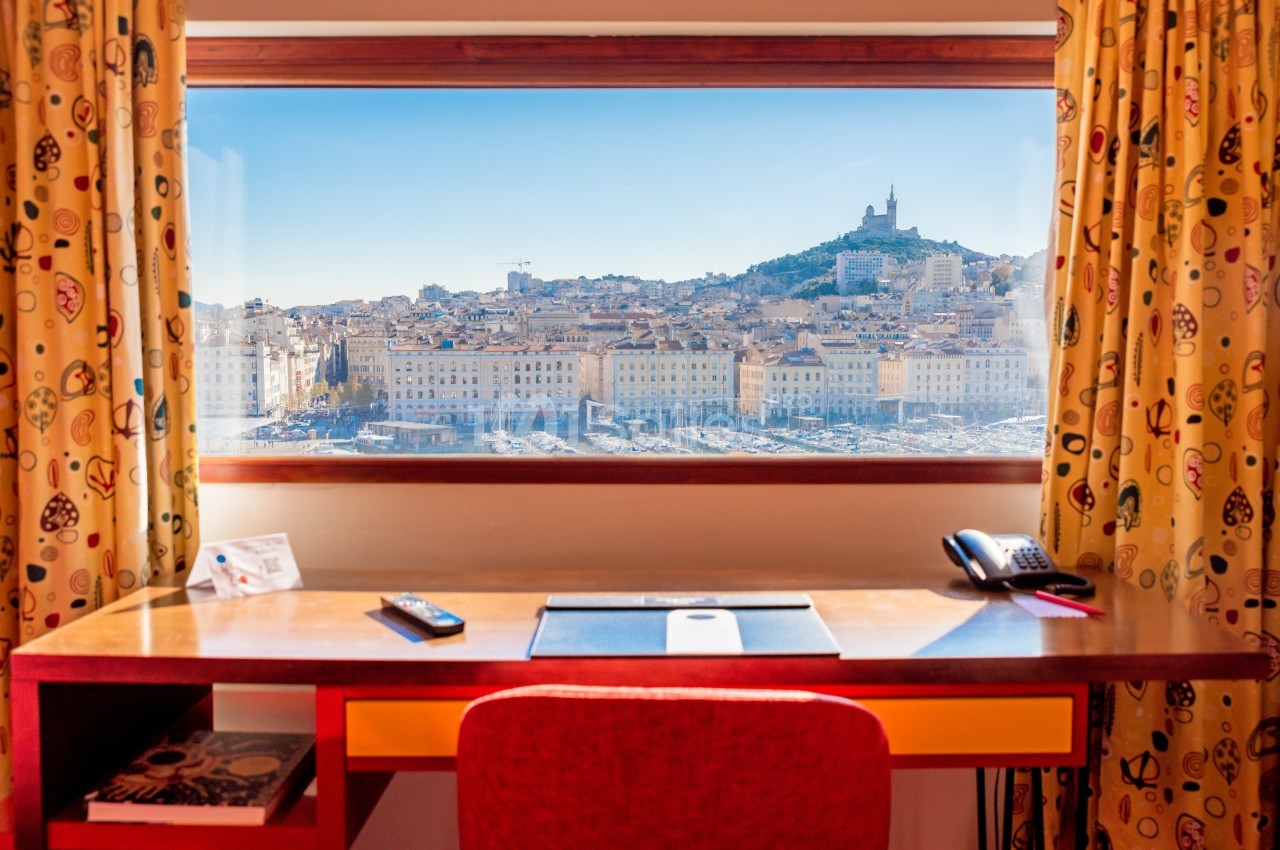Vue sur la ville de Marseille et la basilique Notre-Dame de la Garde depuis une fenêtre encadrée par un bureau.