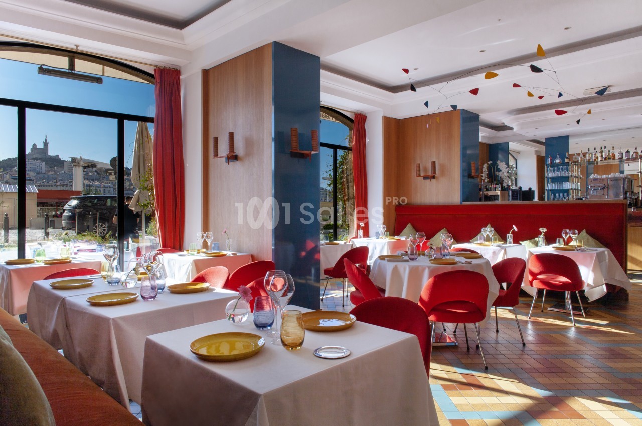 Salle de restaurant lumineuse avec tables dressées, grandes baies vitrées et vue sur un paysage urbain en arrière-plan.
