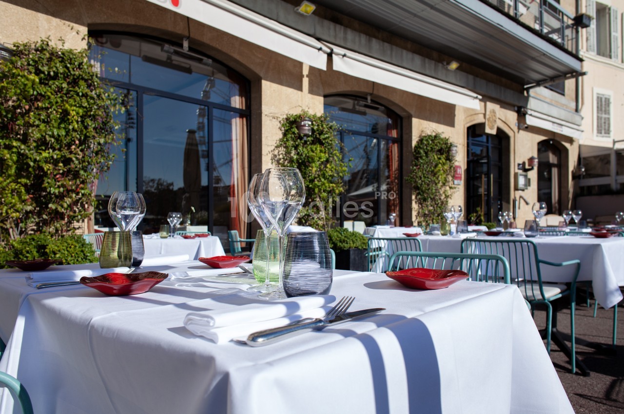 Tables de restaurant dressées en extérieur avec nappes blanches, verres et couverts, devant une façade vitrée.