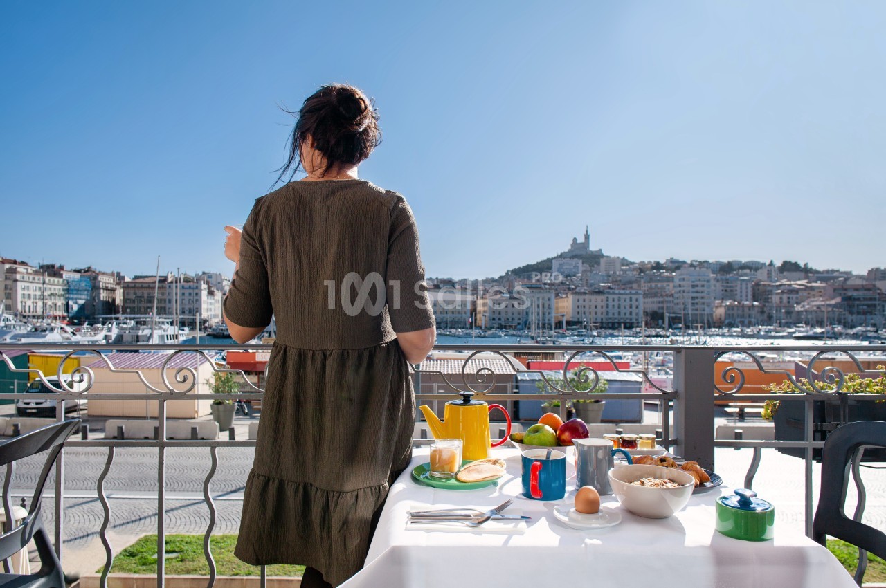 Une femme de dos sur une terrasse, face au Vieux-Port de Marseille, avec une table dressée pour le petit-déjeuner.