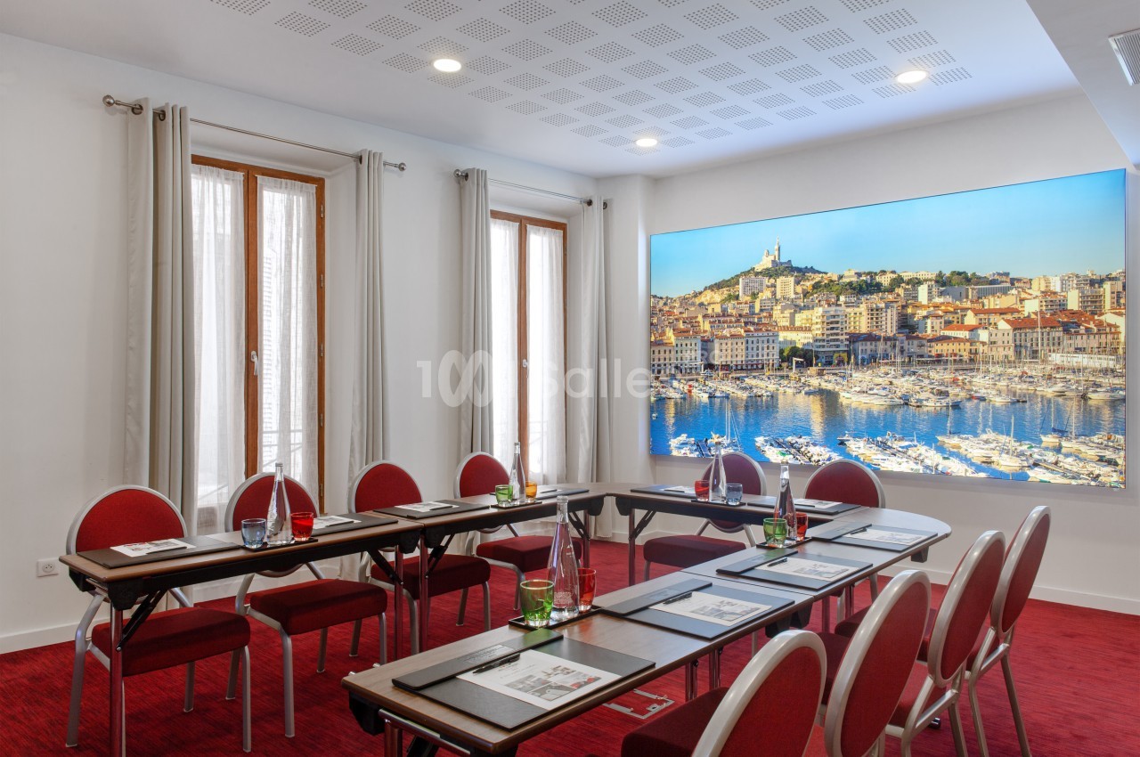 Salle de réunion lumineuse avec tables disposées en U, chaises rouges et vue sur une photo murale de Marseille.
