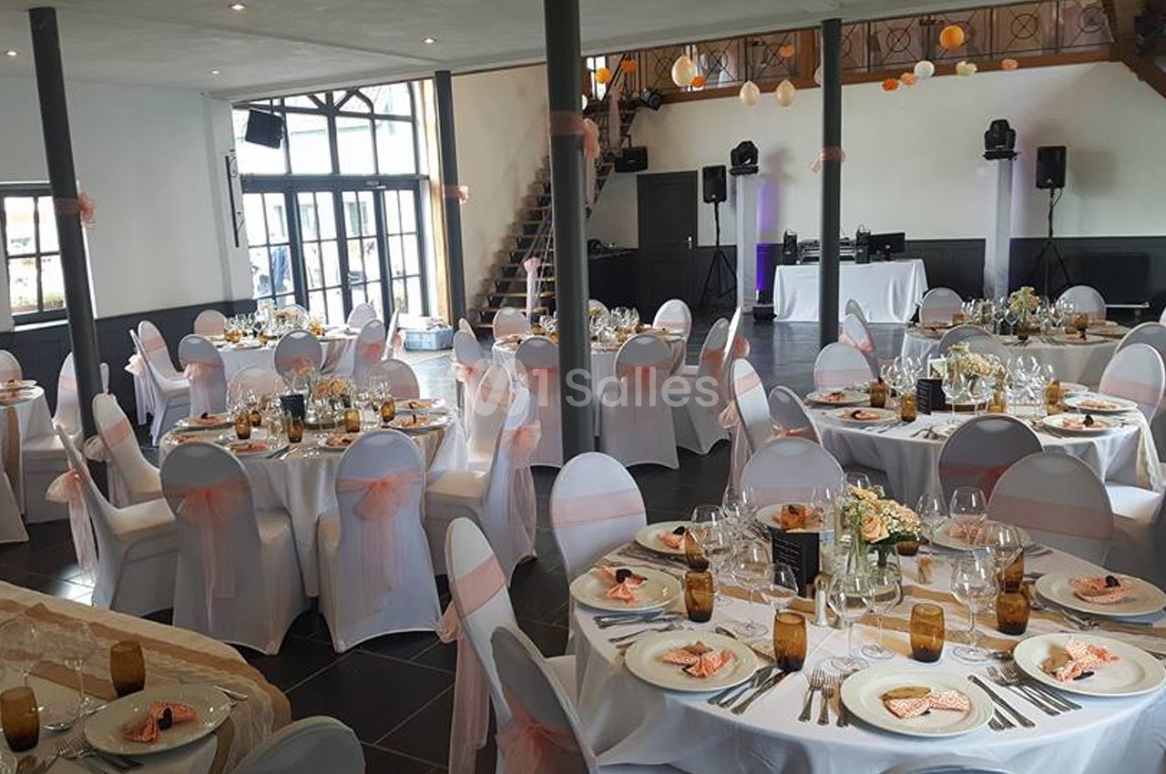Salle de réception décorée avec des tables rondes dressées, nappes blanches, chaises ornées de nœuds roses et lumière…
