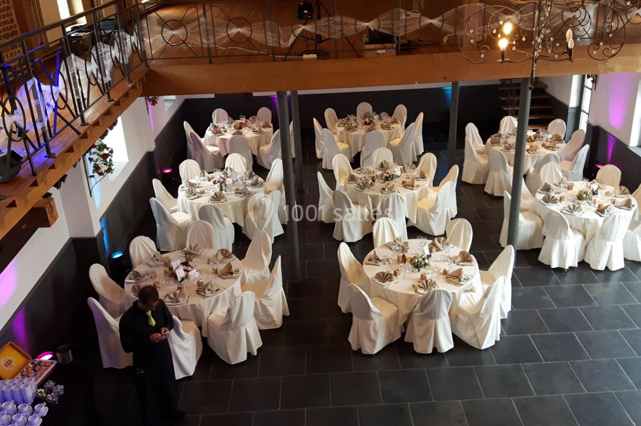 Salle de réception avec tables rondes dressées, nappes blanches et chaises couvertes, vue en plongée.