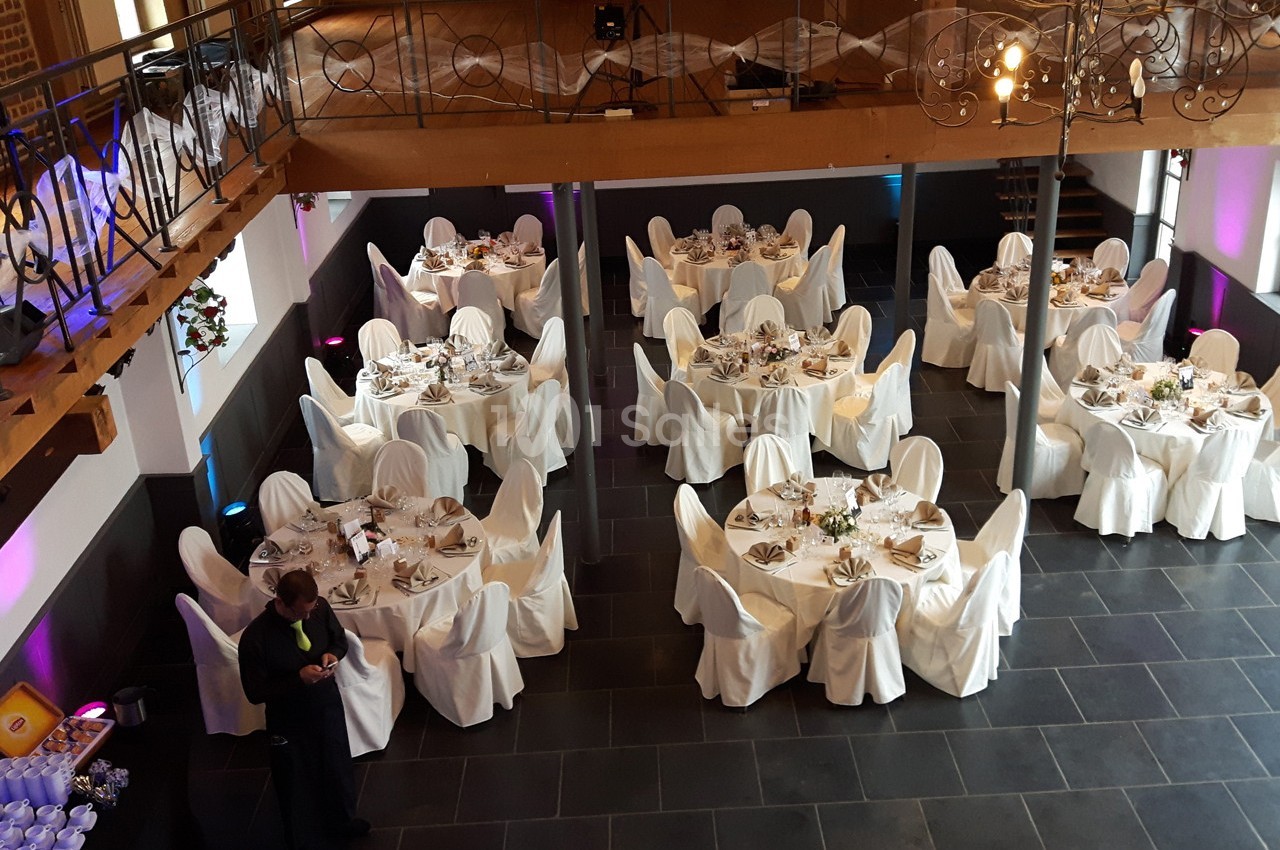 Salle de réception avec tables rondes dressées, nappes blanches et chaises couvertes, vue en plongée.