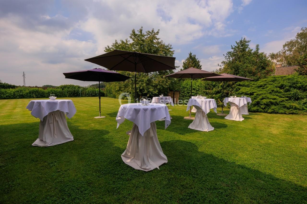 Tables hautes drapées de nappes blanches avec parasols noirs sur une pelouse, entourées de verdure et d'arbres.